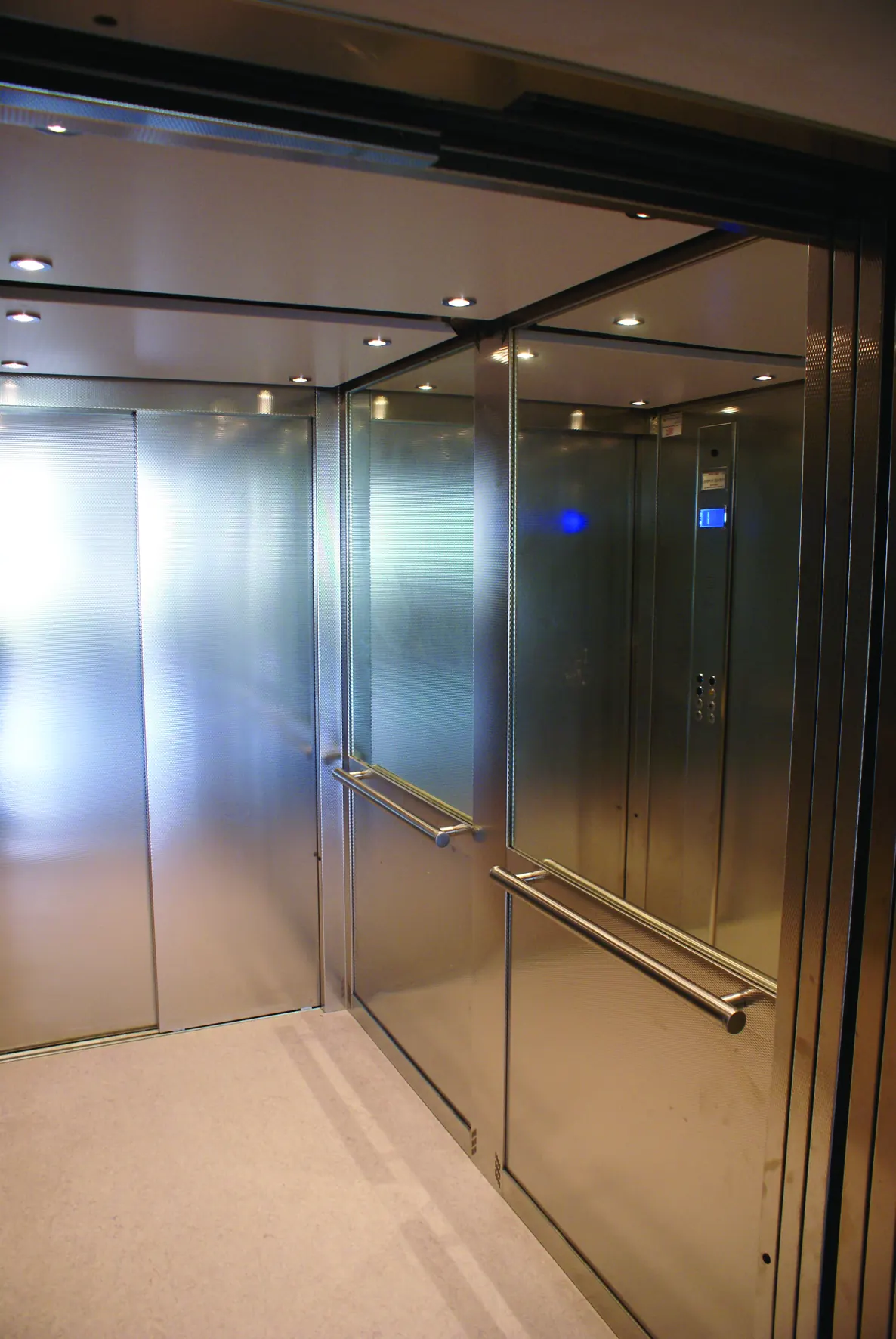 Inside a large lift cabin with brushed stainless steel walls, twin handrails, ceiling spotlights and a control panel