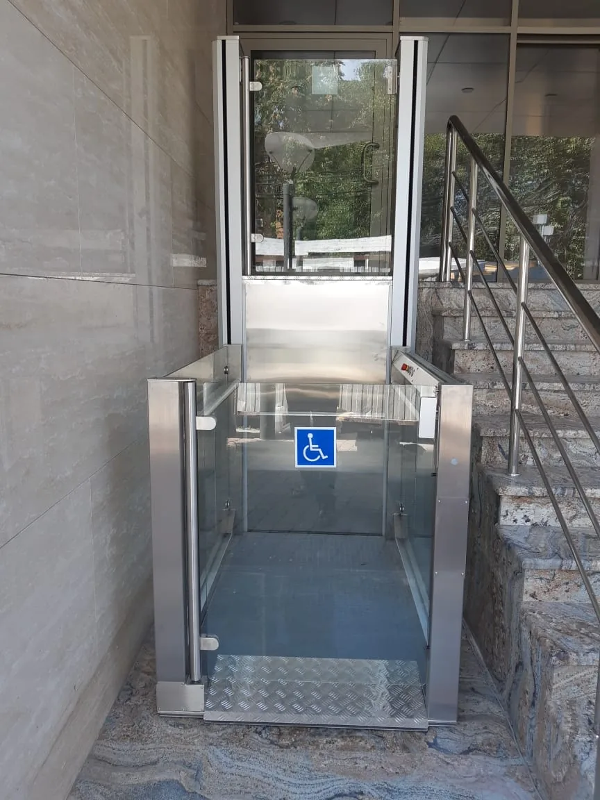 A stainless steel open platform lift with glass gates, a wheelchair sign and a diamond plate floor, at the foot of a marble entrance staircase