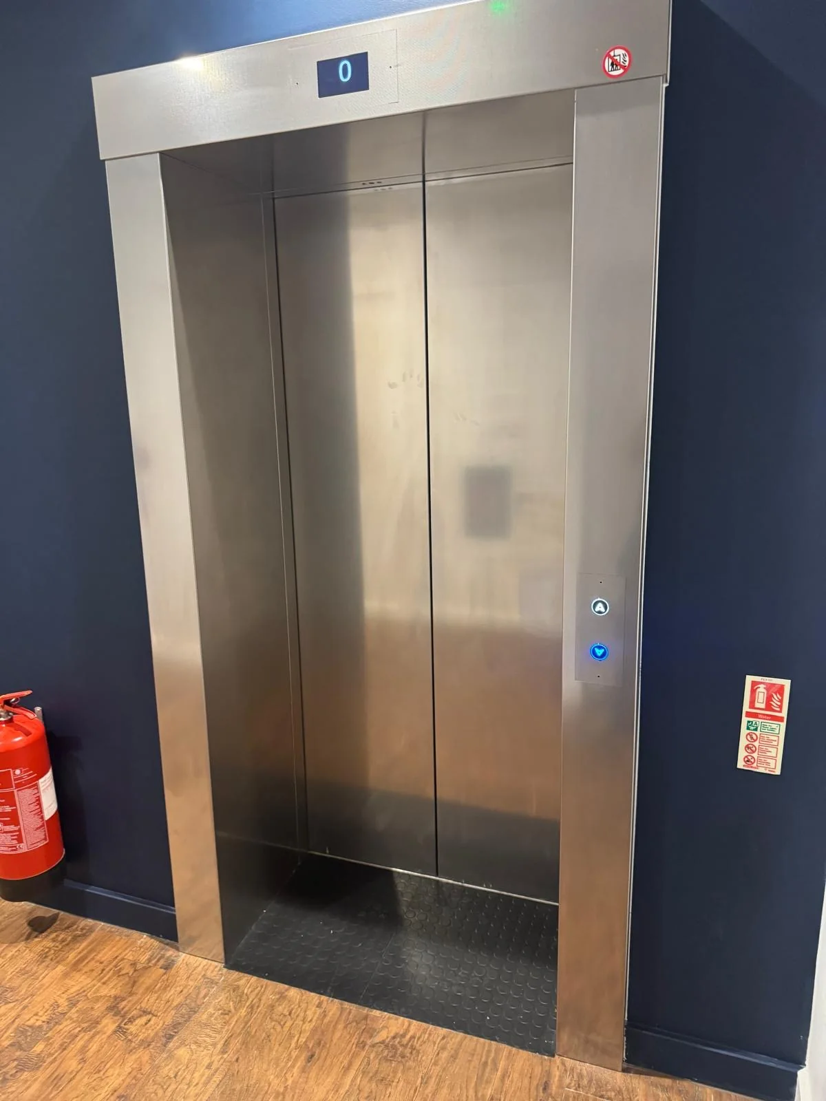 A brushed stainless steel lift entrance set into a navy hotel corridor, with a floor indicator above and call buttons beside it