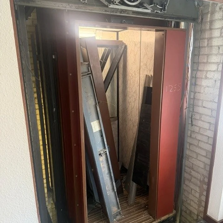 An old lift car being stripped out, door leaves and lining panels stacked in the shaft below the disconnected door operator