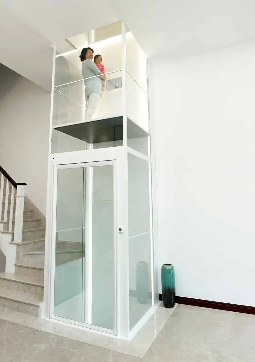 A woman holding a small child rides a white framed glass home lift up beside a staircase