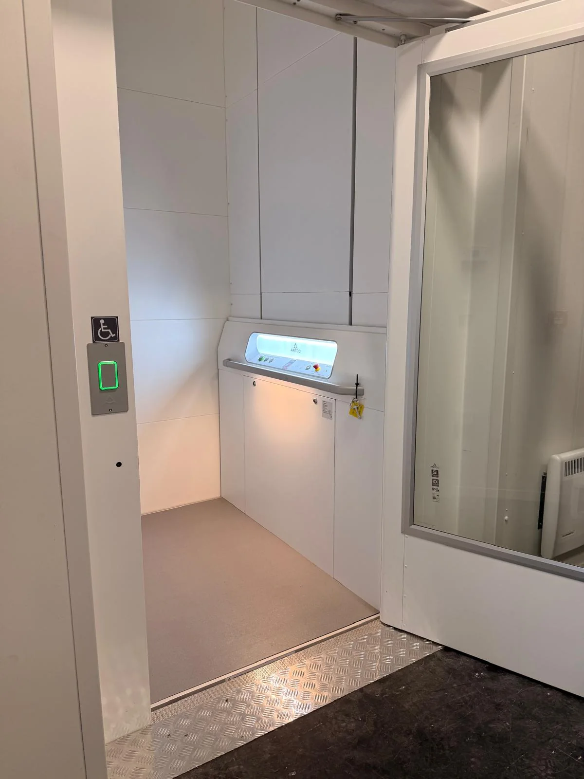 Looking through the open landing door of a platform lift at its platform, the control panel with a handrail, and the call station on the door frame
