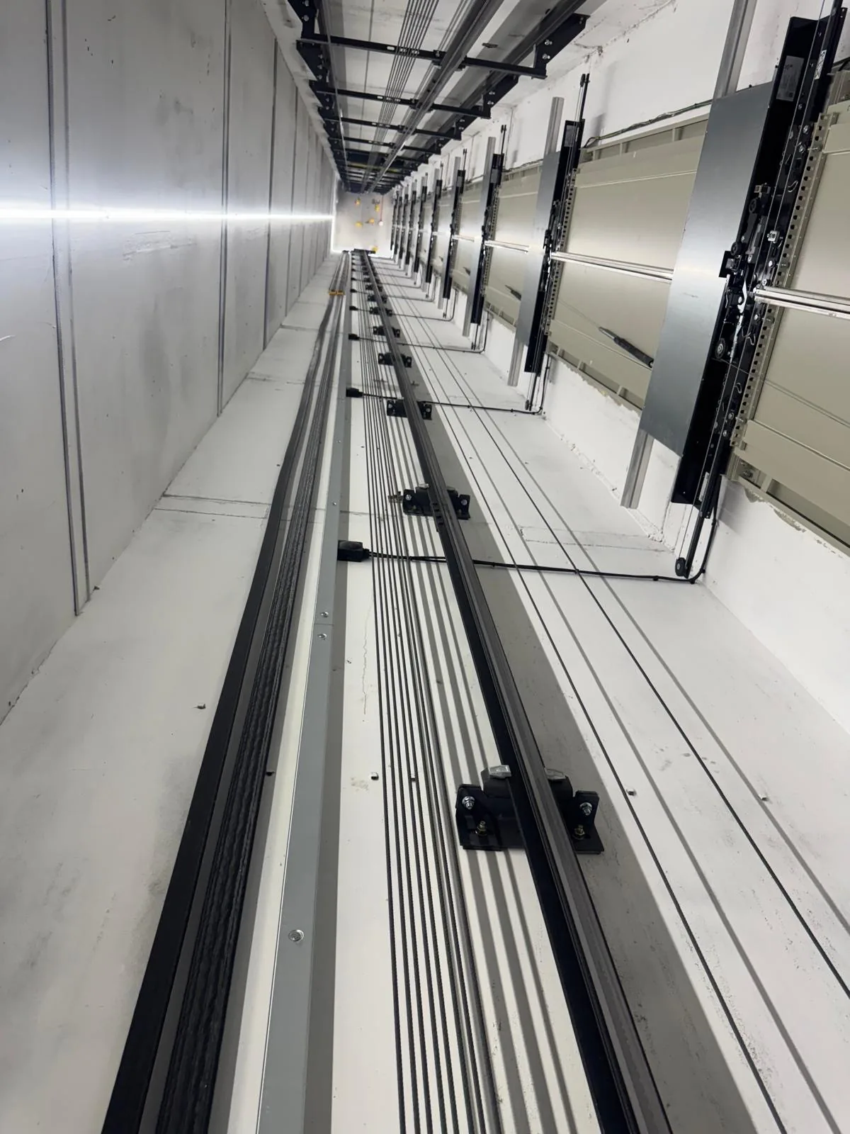 Looking down a freshly painted lift shaft, bright white walls lit by an LED strip, with guide rails and landing door gear running down one side
