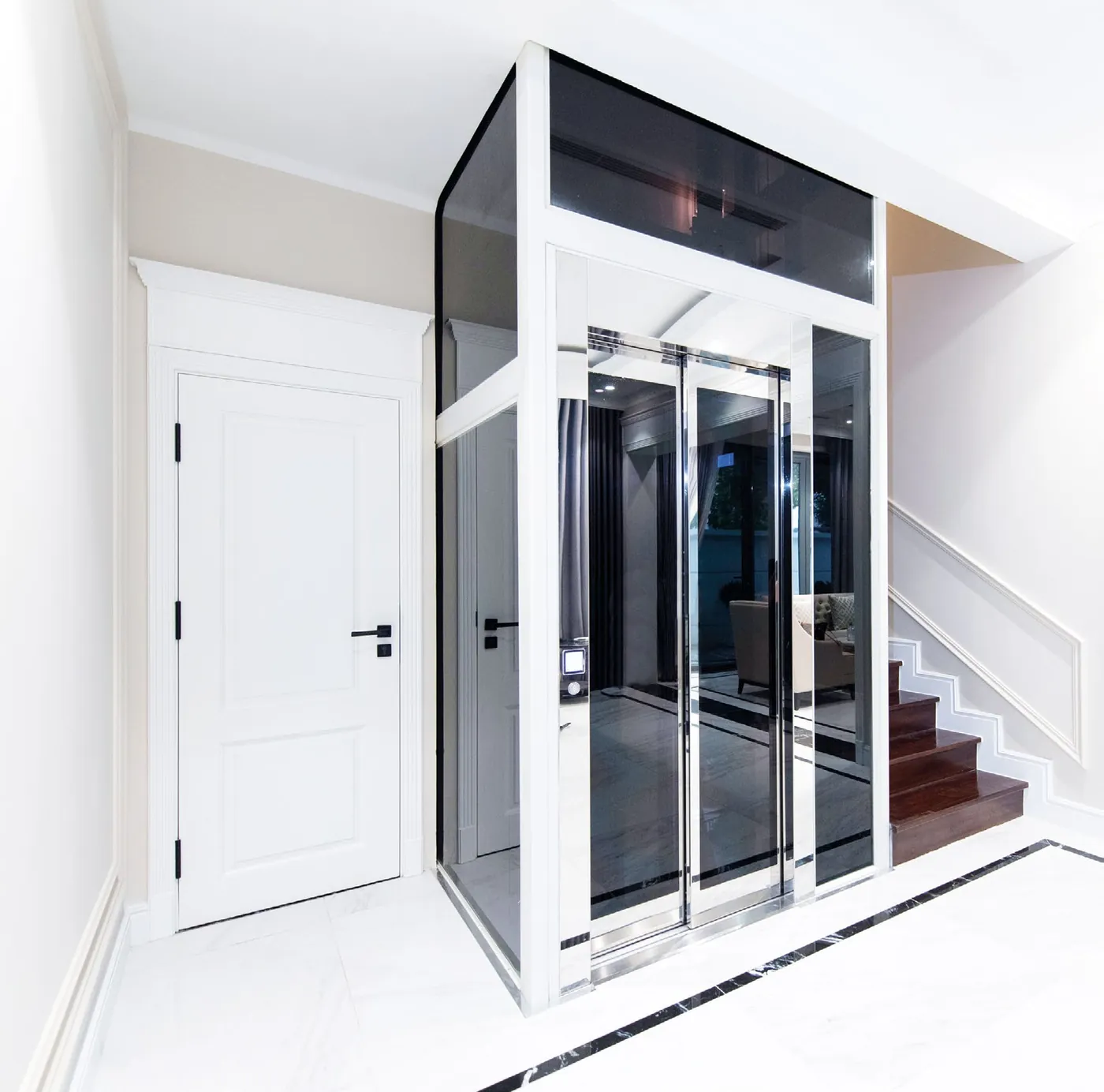 A glazed lift in a white steel structure with tinted glass and sliding doors, in a marble hallway beside a staircase