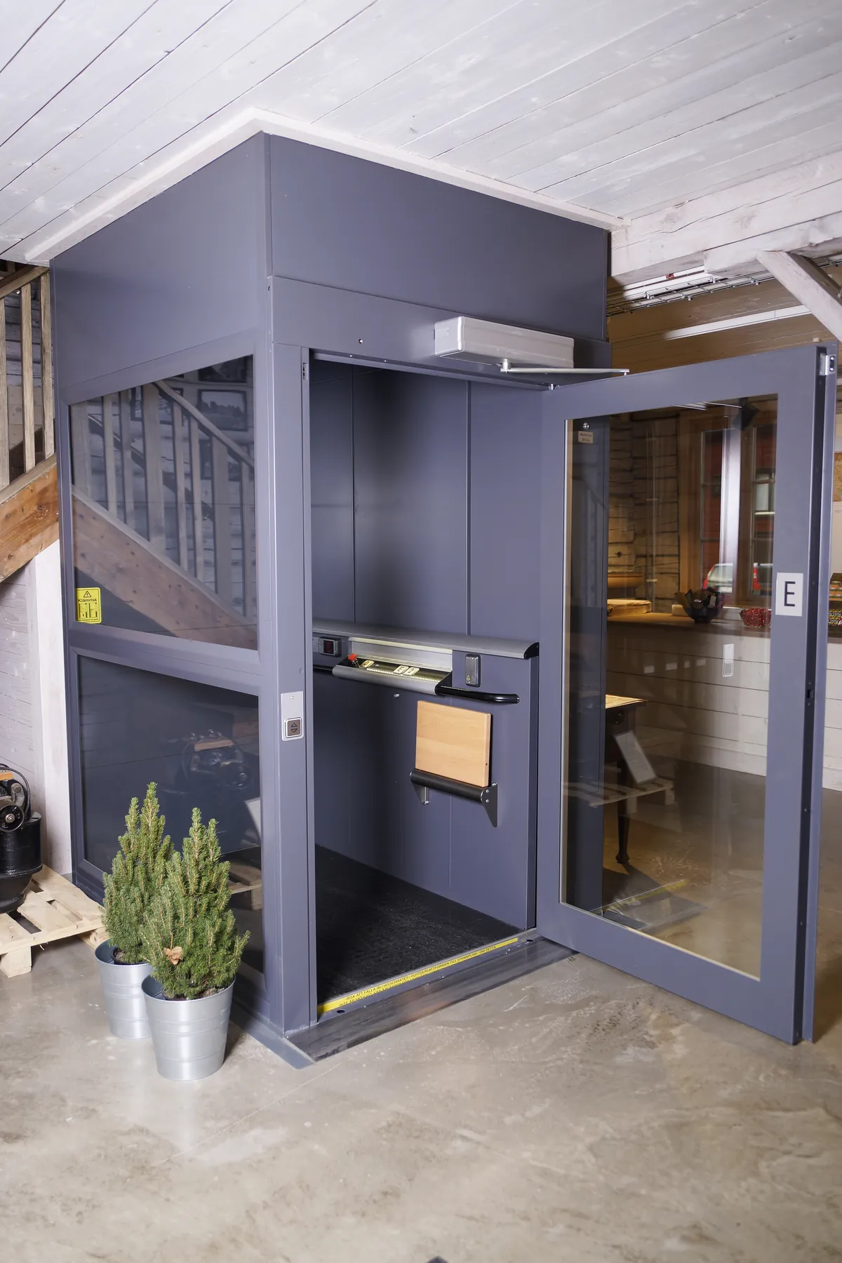 A grey ILS 05K platform lift with its glazed swing door open, showing the platform, control rail and fold-down seat