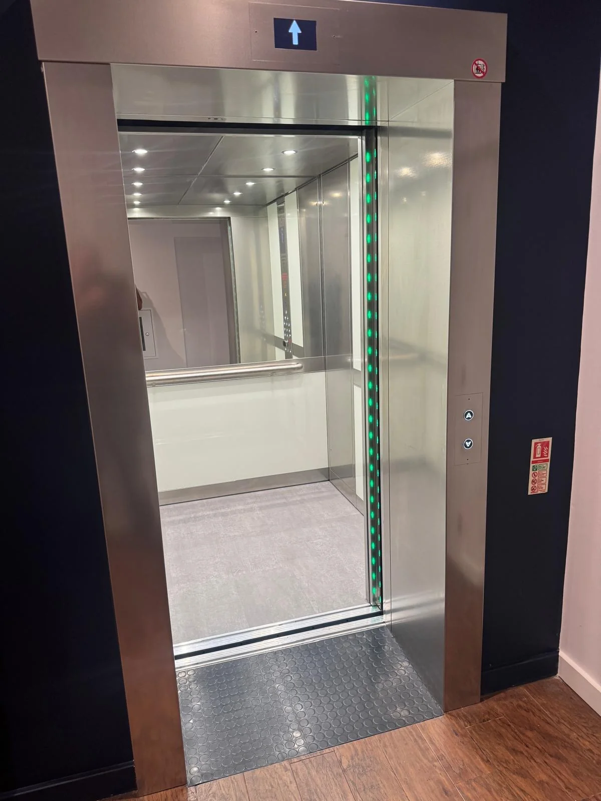 The new lift at a landing with its stainless steel doors open, showing the mirrored car and a green-lit light curtain down the door edge