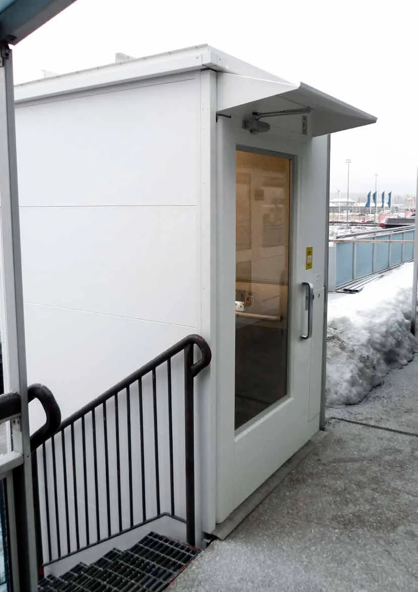 A white outdoor Aritco HomeLift Access with a canopy over its glazed door, at the top of an outside staircase with snow on the ground