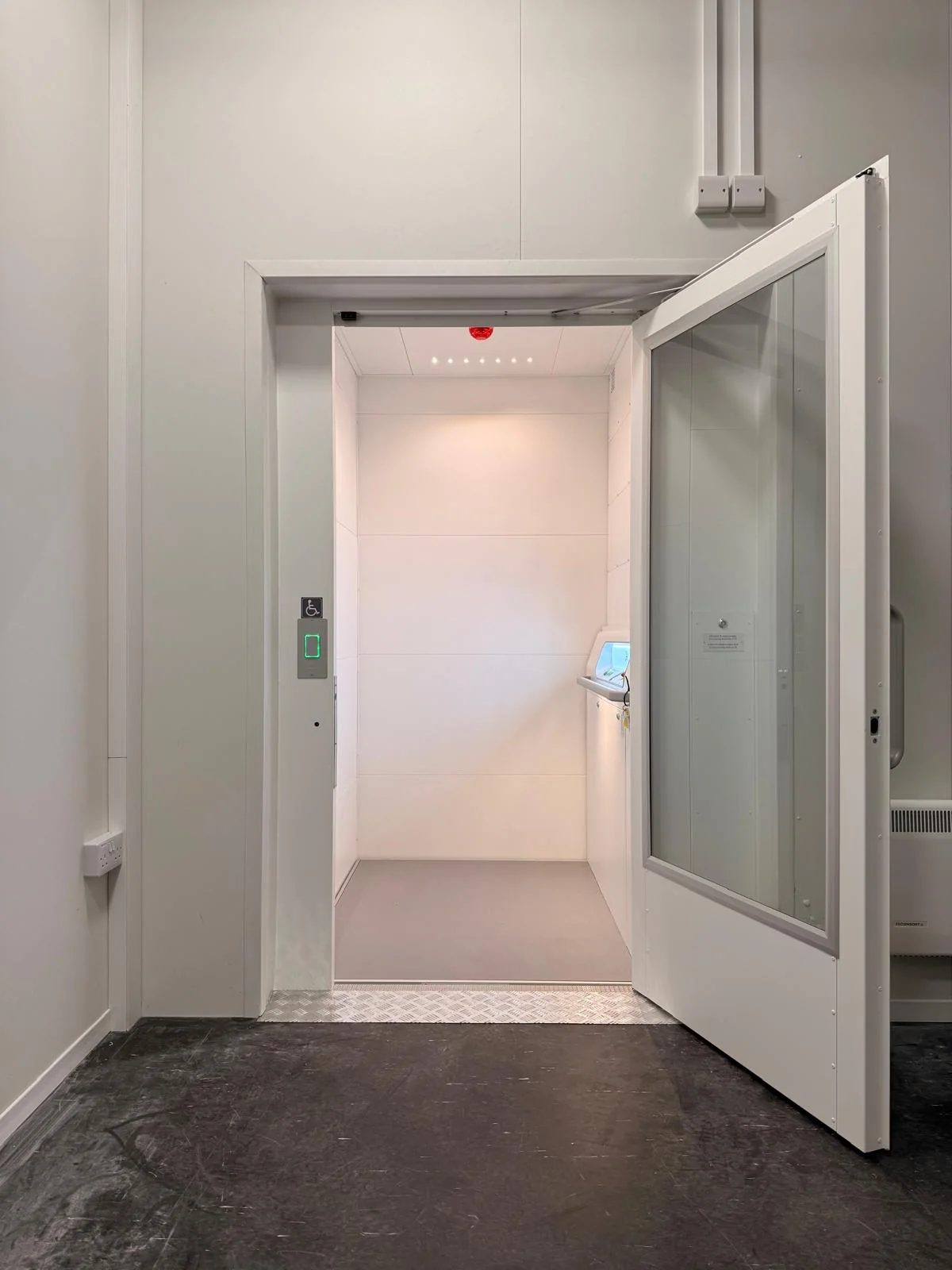 A white lift landing door with a large vision panel swung open onto a bright cabin, with a level chequer plate threshold and a call station beside it