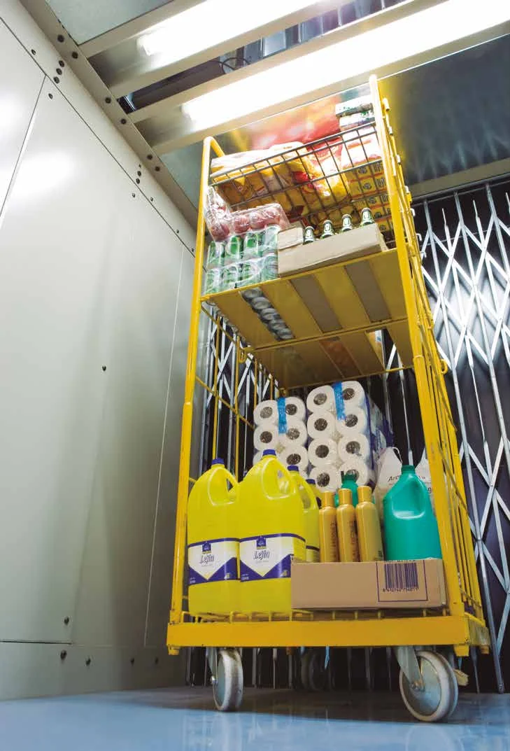 A yellow roll cage of groceries and cleaning supplies inside a goods lift car with beige steel walls and folding gates